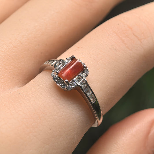 Angled view of a red-toned rectangular jadeite ring worn on a finger, showing the silver setting and clear accent stones.