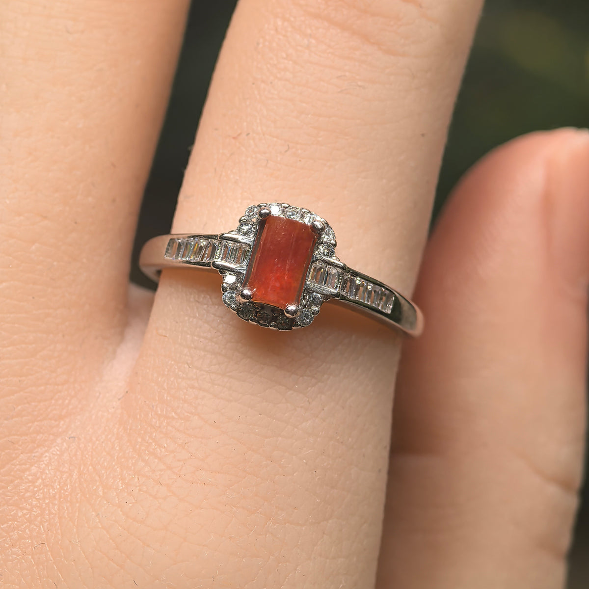 Front view of a red jadeite ring worn on a finger, with a clear halo and rectangular accents along both shoulders.