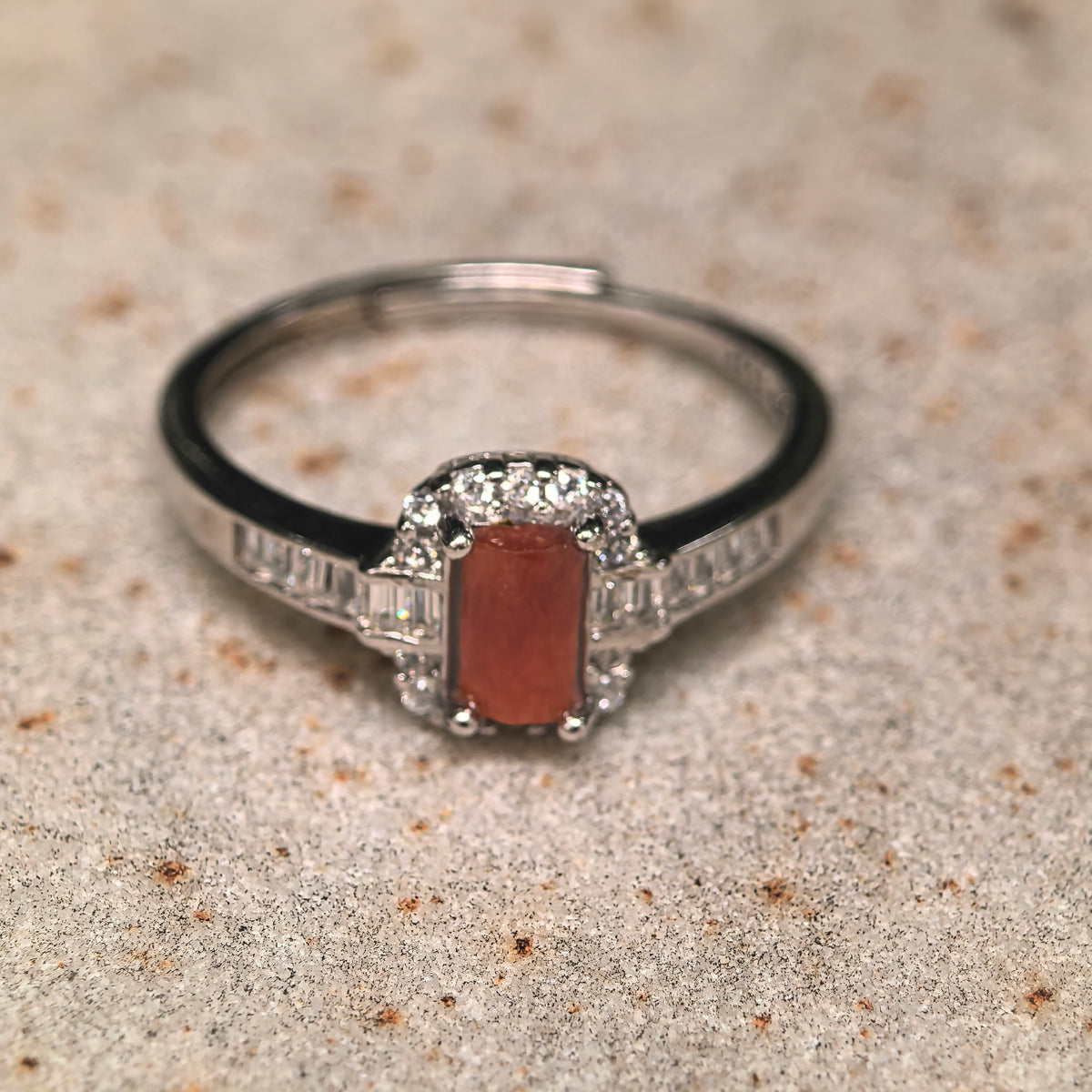 Front view of a red-toned rectangular jadeite ring on a textured stone surface, showing the clear halo and silver band.