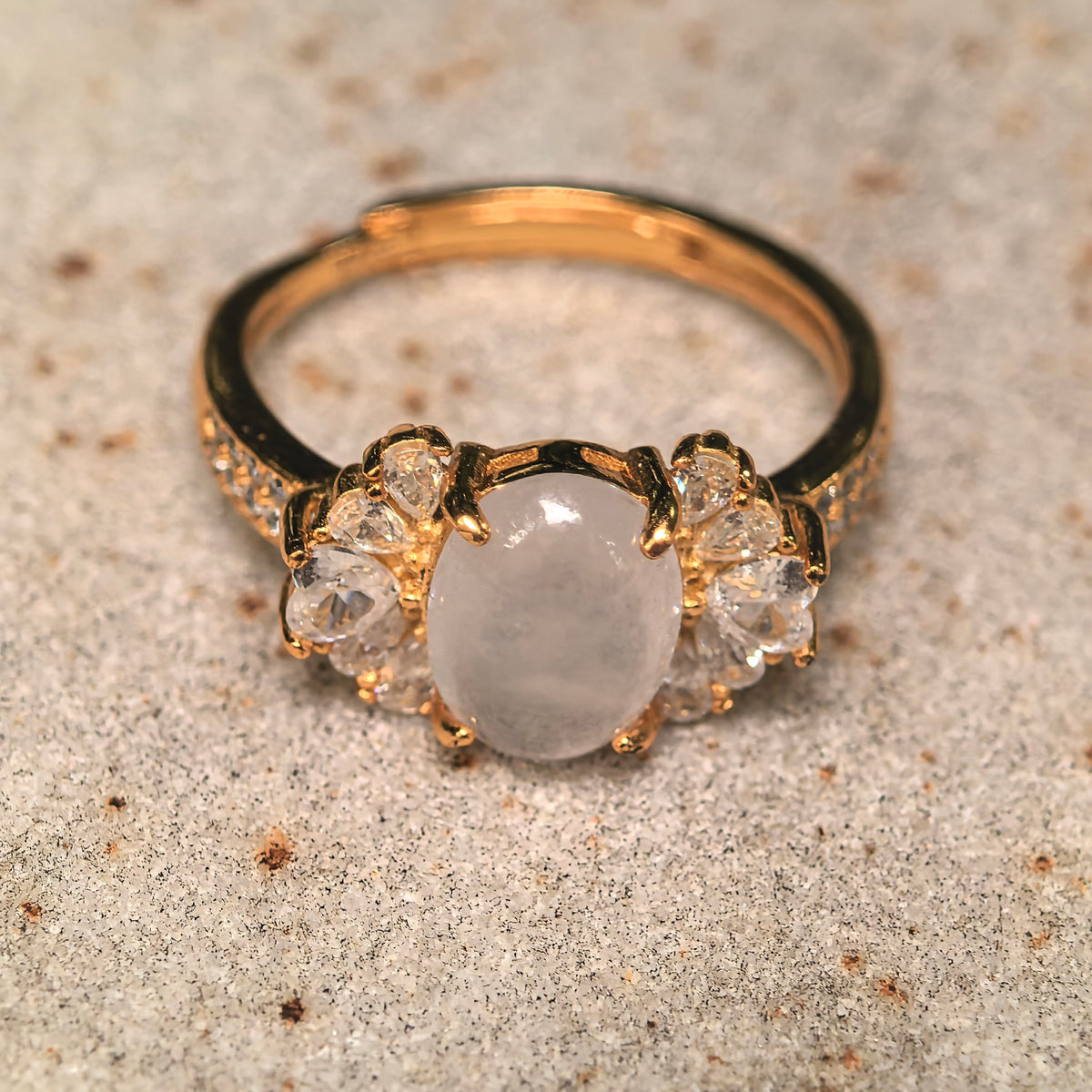 White oval jadeite ring on a textured stone surface, showing clear side clusters and an open gold-tone silver band.