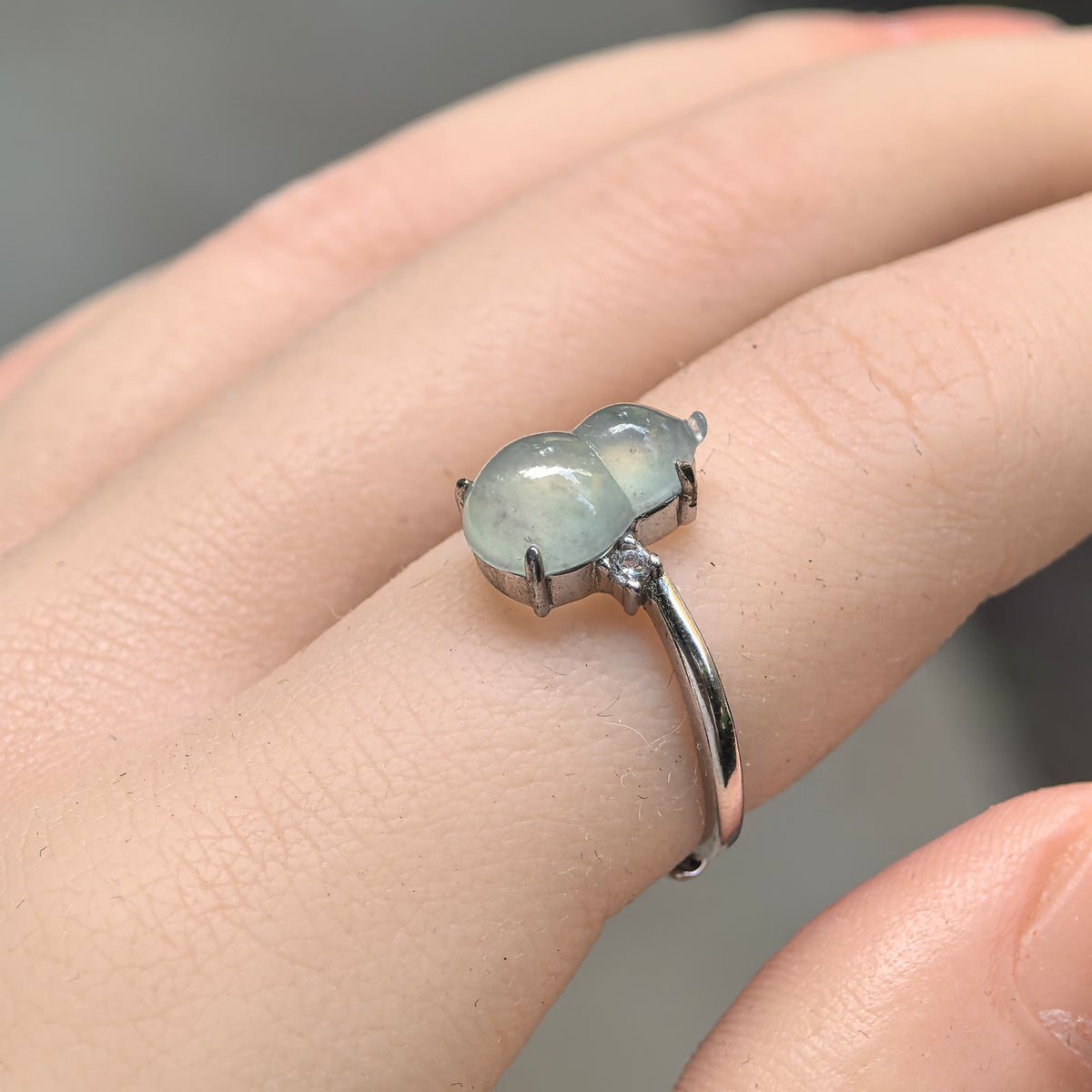 Angled view of a pale green jadeite gourd ring worn on a finger, showing the polished carving, silver prongs and a clear side accent stone.