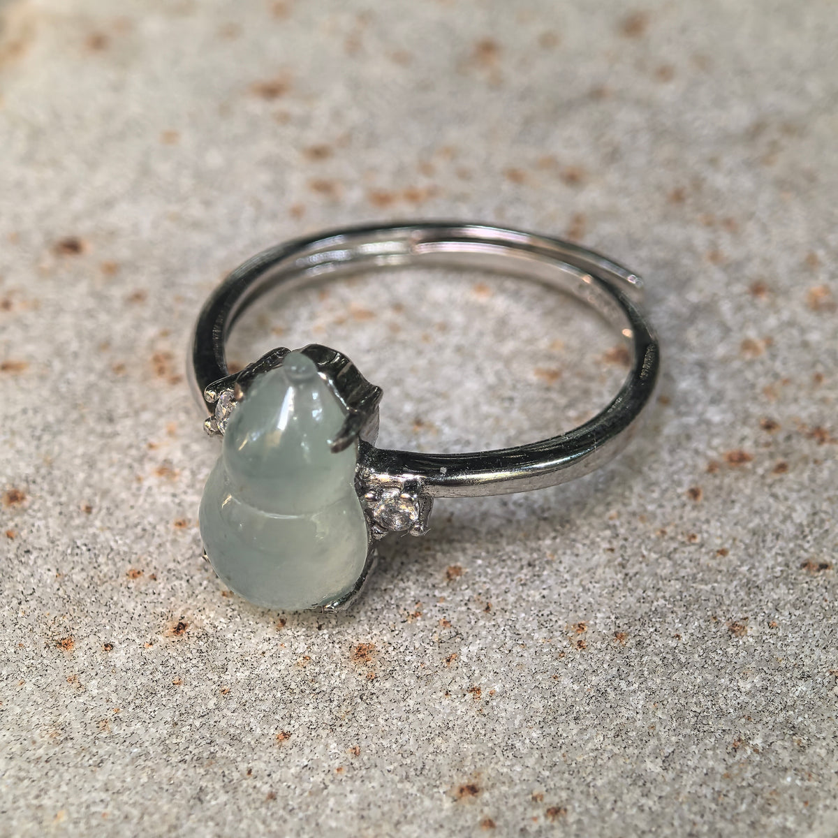 Angled view of a pale green jadeite gourd ring on a stone surface, showing the prong setting, side accents and overlapping adjustable ring band.