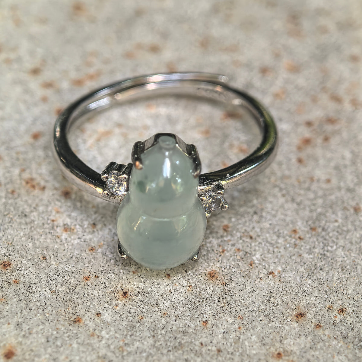 Pale green jadeite gourd ring on a textured stone surface, showing the upright carving, small stem and clear accent stones on both sides.