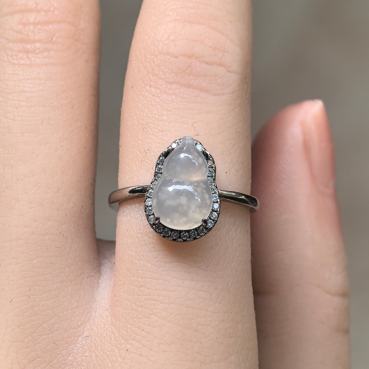 Front view of a white jadeite gourd ring worn on a finger, showing snowflake-like inclusions within the carving and a halo of clear accent stones.