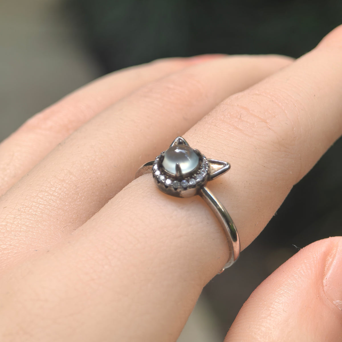 Angled view of an icy white jadeite cat ring worn on a finger, showing the round cabochon, clear accent halo and pointed silver ears.