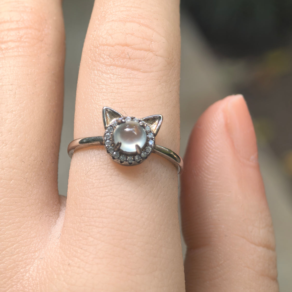 Front view of the white jadeite cat ring on a finger, with two triangular silver ears above the round cabochon and clear accent halo.