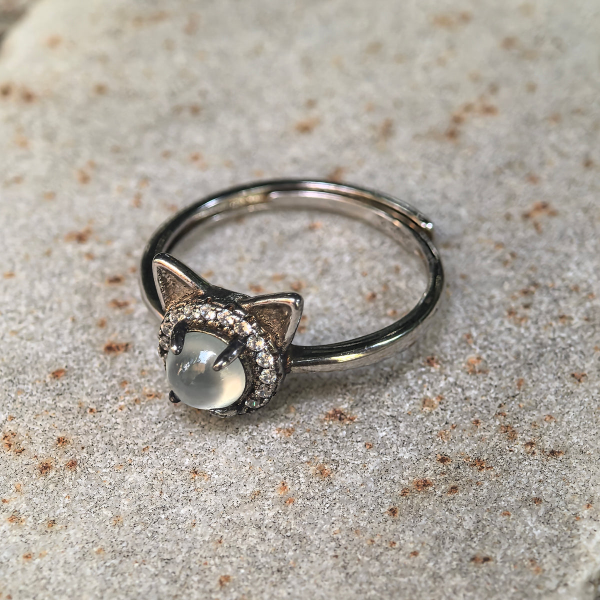 White jadeite cat ring resting at an angle on stone, showing the S925 silver setting, clear accent halo and open adjustable ring band.