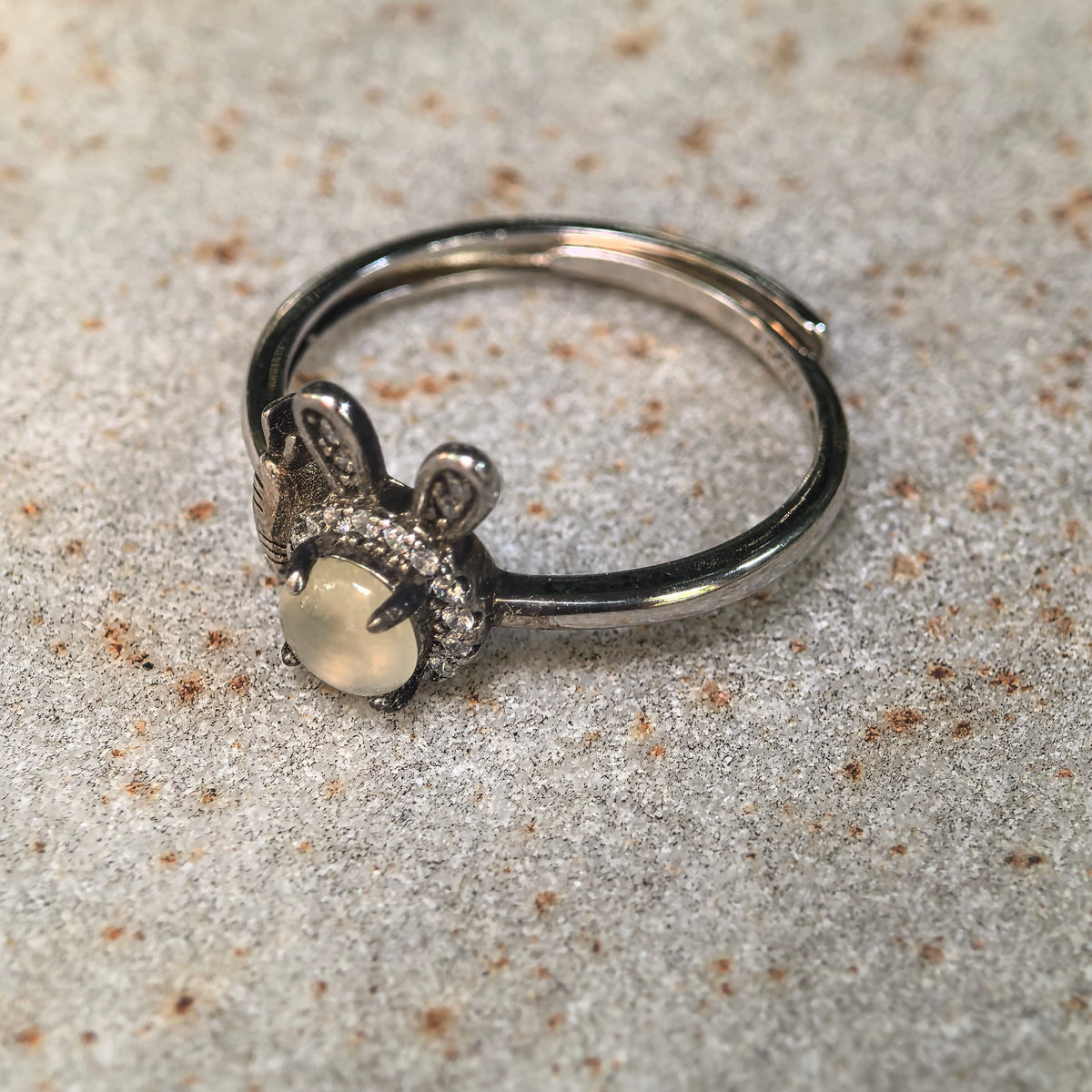 White jadeite rabbit ring resting on a stone surface, showing the round cabochon, carrot detail and open S925 silver ring band.