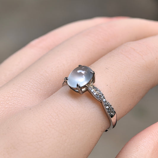 Angled close-up of a white oval jadeite ring worn on the finger, showing its translucent cabochon, silver prongs and intertwined shoulders with clear accent stones.