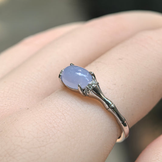 Angled close-up of a purple oval jadeite ring worn on the finger, showing its silver prongs, clear side accents and tapered segmented band.