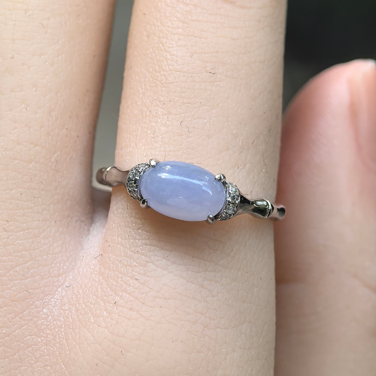Front view of a purple jadeite ring on the finger, with a horizontally set oval cabochon and clear accent stones on both sides.
