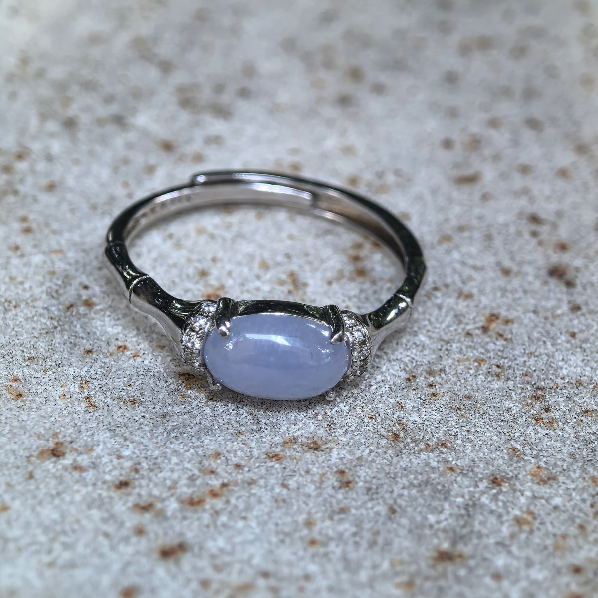 Purple oval jadeite ring displayed on textured stone, showing the four-prong silver setting, clear side accents and open ring band.