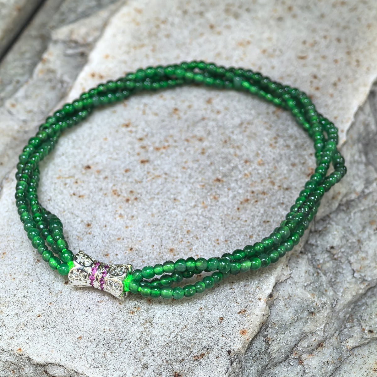 Green jadeite bead bracelet arranged on textured stone, with a silver-tone bow-shaped connector featuring clear and pink accents.