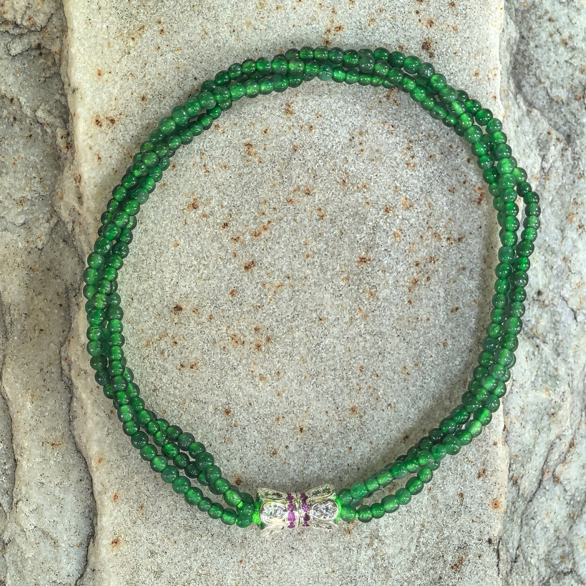 Overhead view of a green jadeite bracelet on stone, showing the complete twisted bead design and decorative bow connector.