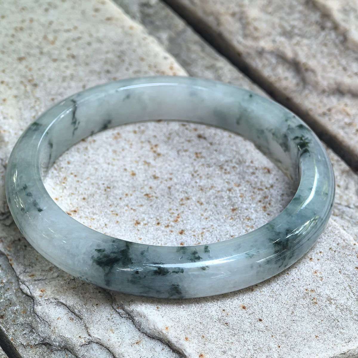 Angled view of a jadeite D-shape bangle on textured stone, showing the rounded outer surface and scattered dark green patterns.