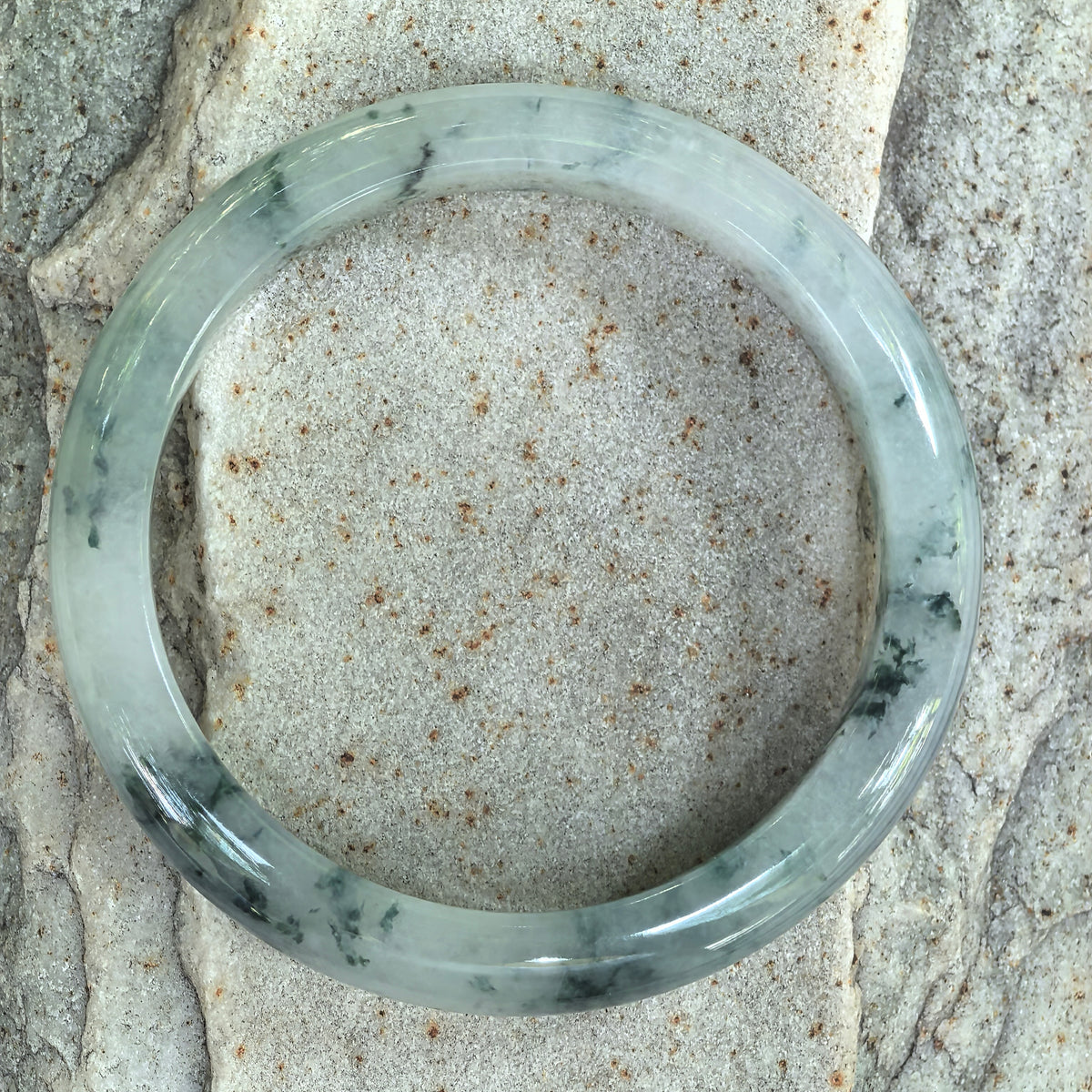 Overhead view of a jadeite bangle on stone, showing its circular outline and dark green floating patterns around the white base.