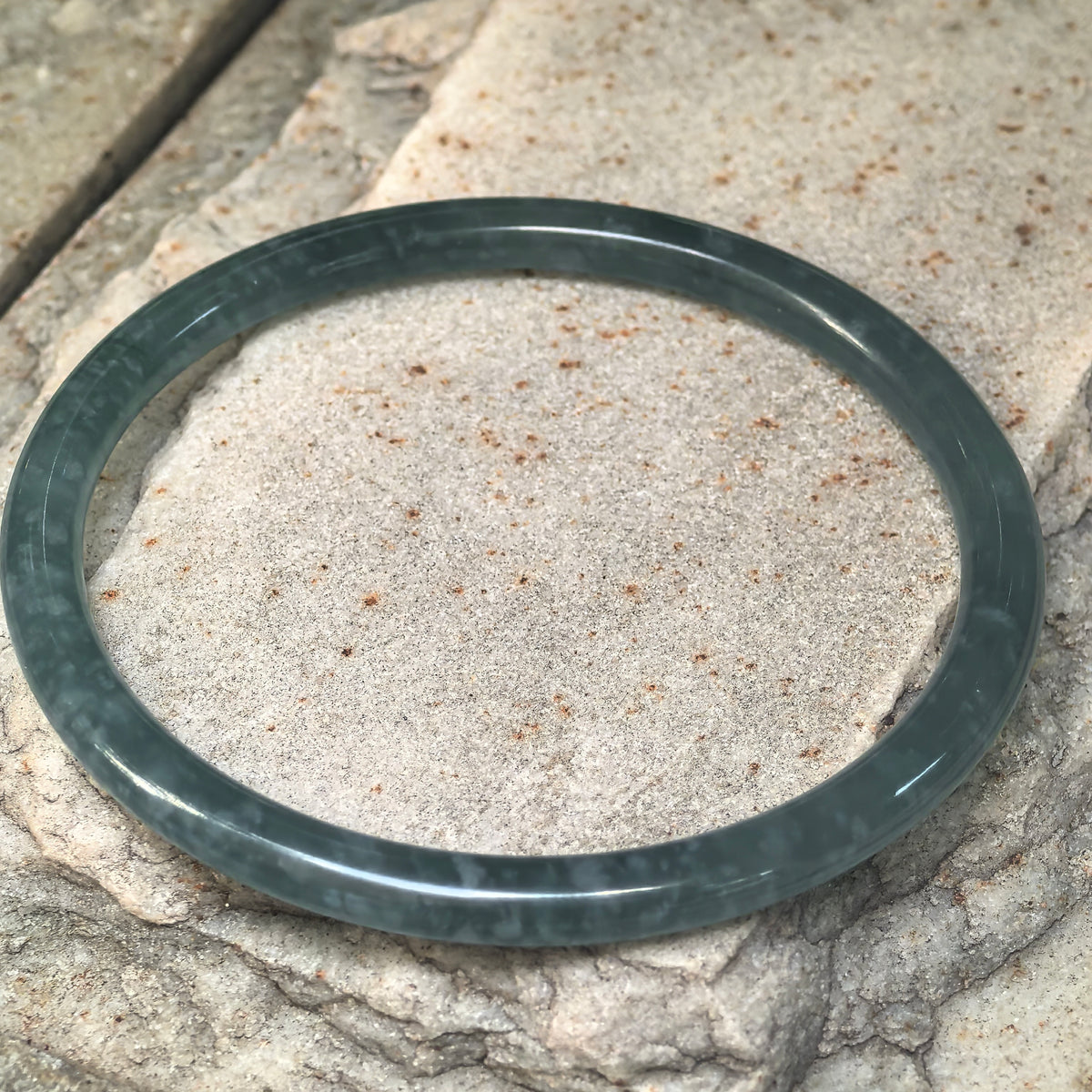 Angled view of a slim green jadeite bangle resting on a textured stone surface, showing its rounded edges and internal cotton-like patches.