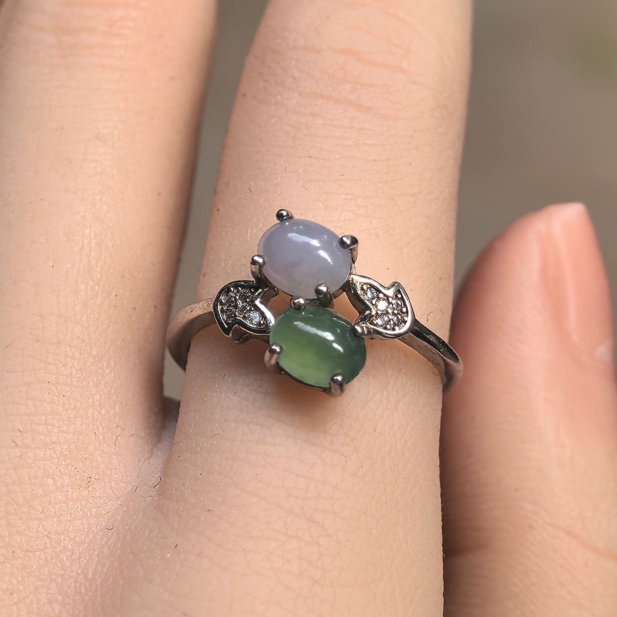 Front view of a purple and green jadeite ring on a finger, with offset oval cabochons and curved silver shoulder motifs.