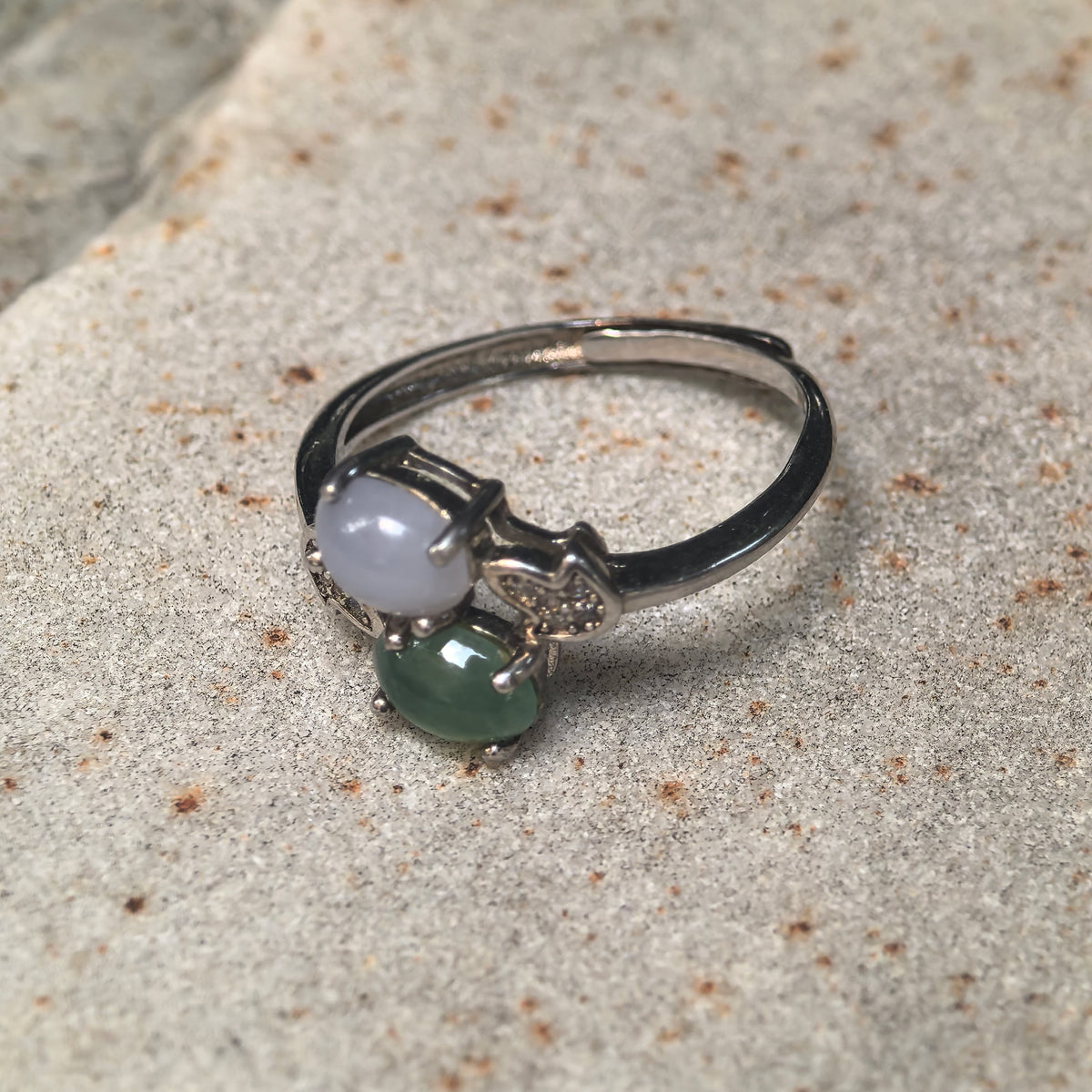 Purple and green twin-stone jadeite ring on a textured stone surface, showing the side settings and overlapping ends of the open silver ring band.