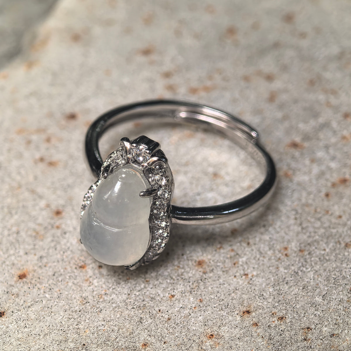 Angled view of a white jadeite gourd ring on a stone surface, showing the halo setting, top accent and overlapping adjustable ring band.