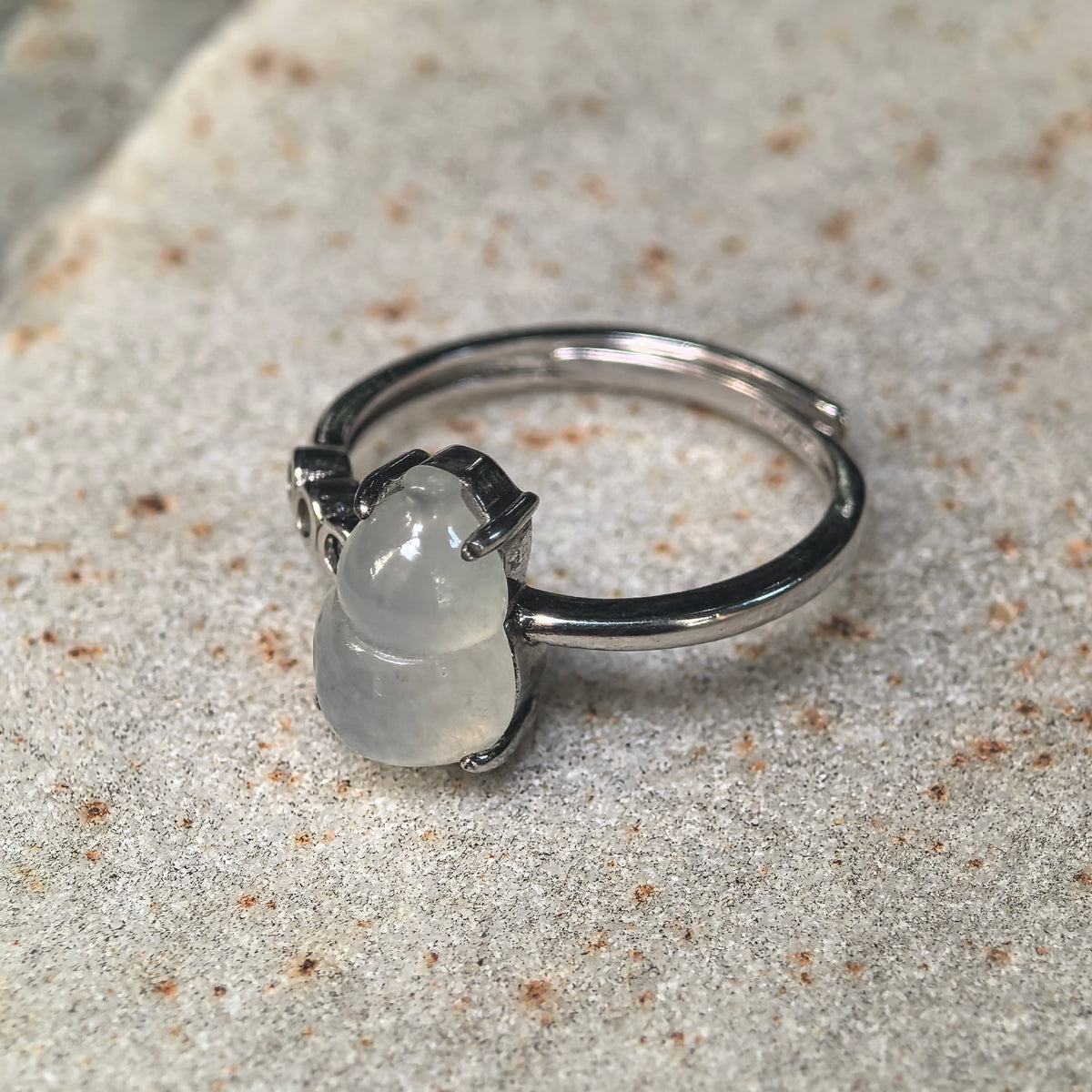 Angled view of a white jadeite gourd ring on a stone surface, showing the raised prong setting, polished silver ring band and overlapping adjustable opening.