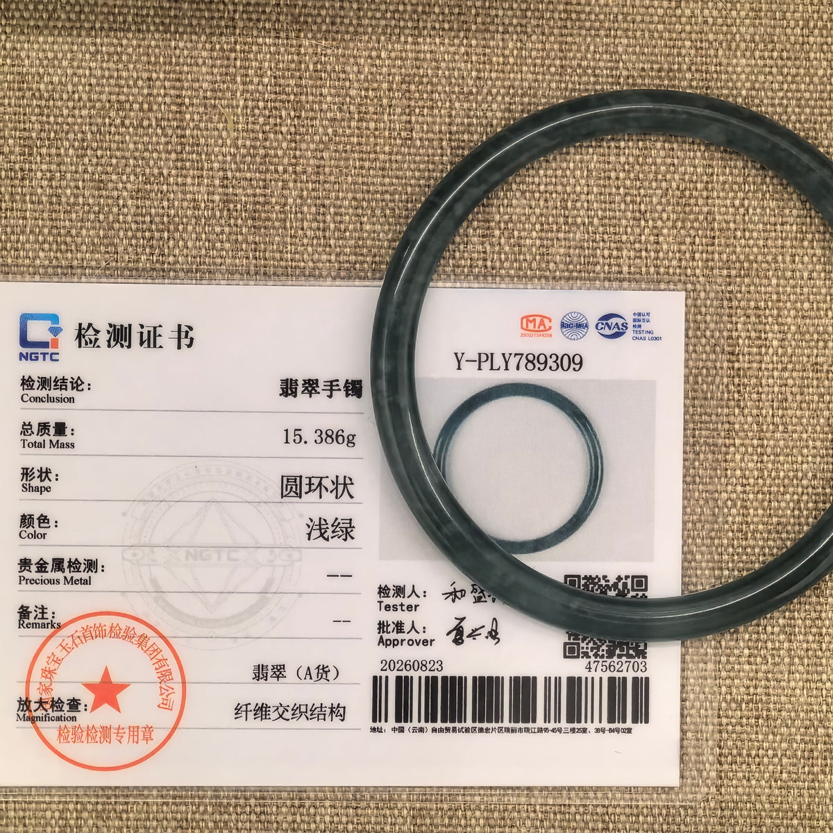 Green jadeite bangle placed over its matching NGTC certificate on a woven fabric background.