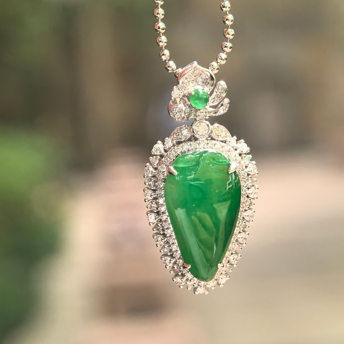 Bright green pear-shaped jadeite pendant suspended from a display chain, showing its diamond border and decorative 18K gold upper setting.