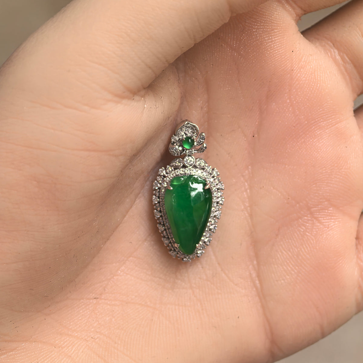 Green jadeite pendant resting in an open palm, showing its pear-shaped centerpiece and surrounding diamond setting.