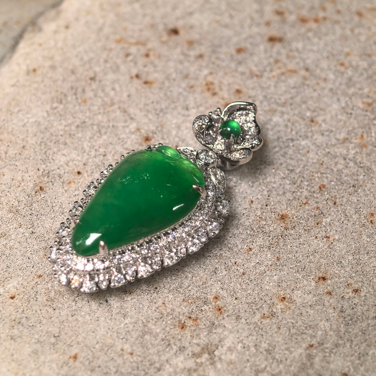 Angled close-up of a bright green jadeite pendant on stone, showing carved leaf details and its 18K gold setting with diamonds.