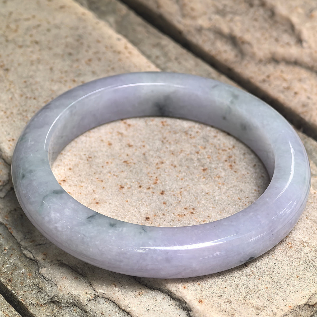 Pale lavender jadeite D-shape bangle resting at an angle on stone, showing scattered dark patterns and its flat inner surface.