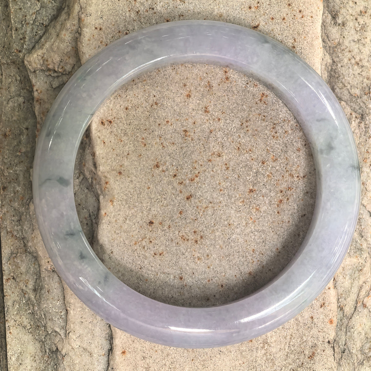Overhead view of a pale lavender jadeite bangle on stone, showing lighter sections and irregular dark markings around the band.