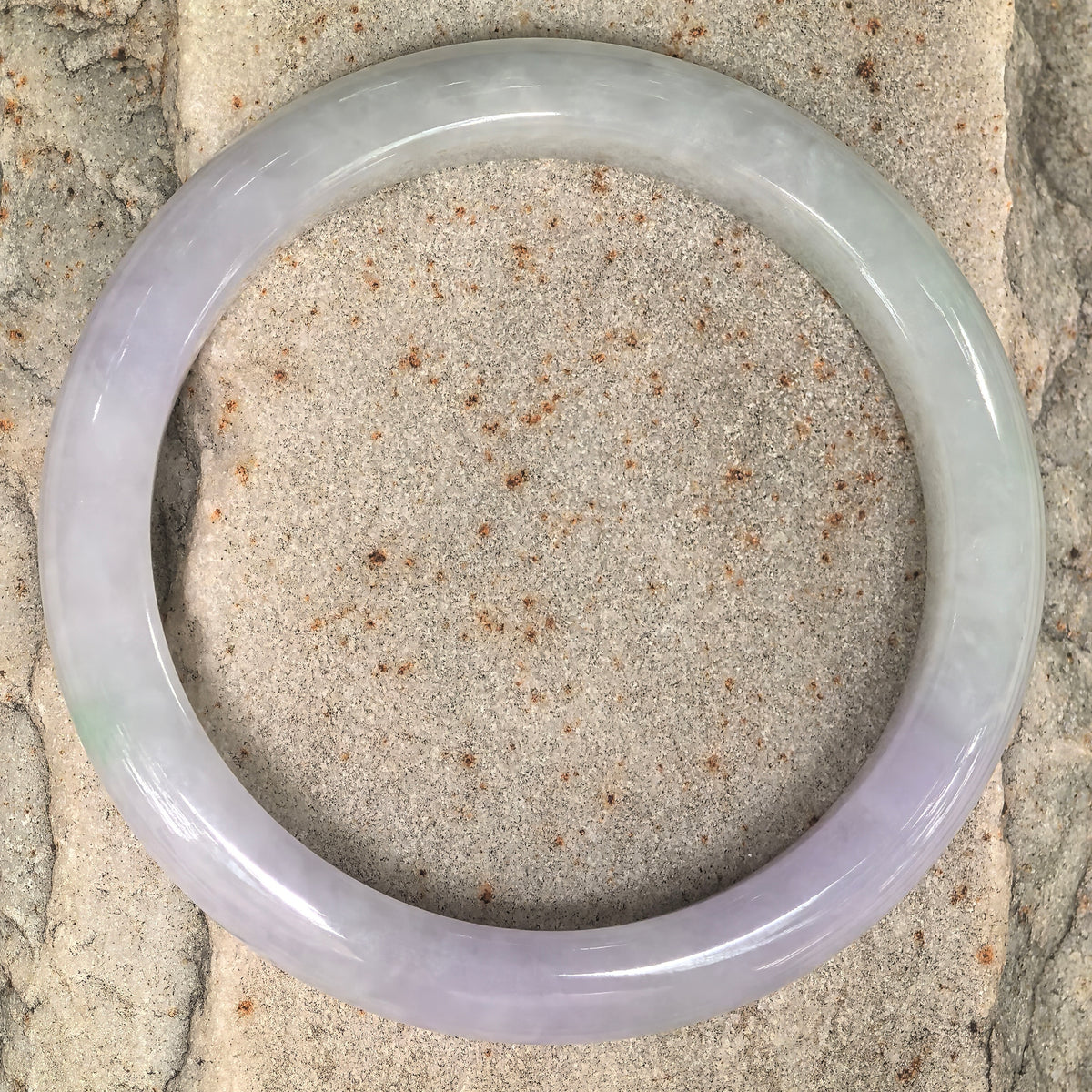 Overhead view of a pale lavender and light green D-shape jadeite bangle on a textured stone surface.