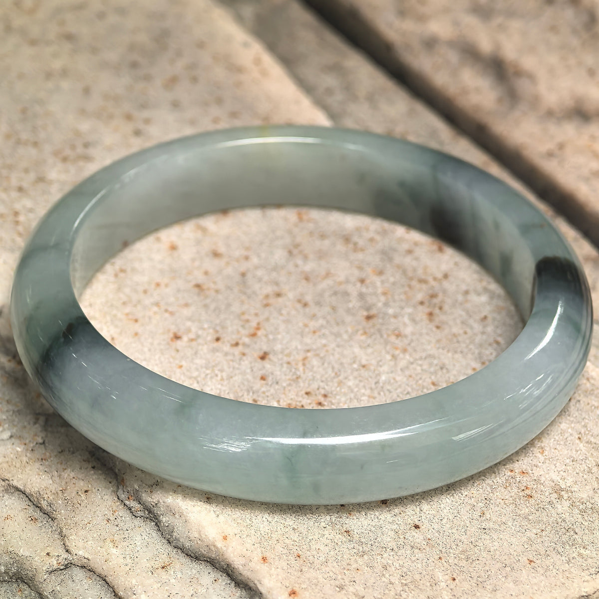 Angled view of a light green D-shape jadeite bangle on stone, showing dark floating patterns and variations in translucency.