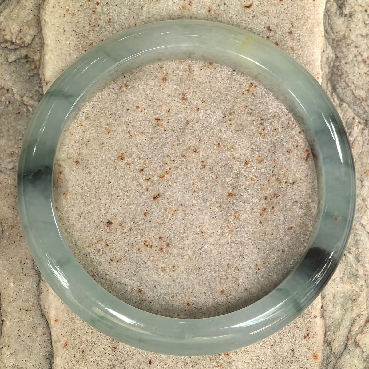 Overhead view of an icy glutinous jadeite bangle on stone, with a pale green base, dark patterns and small inclusion spots.