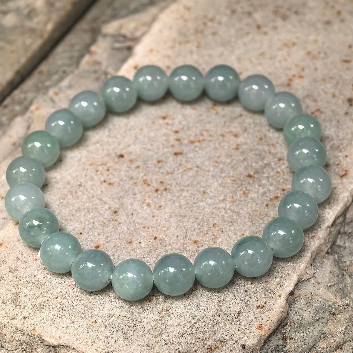 Angled view of a light green round bead jadeite bracelet resting on a textured stone surface.