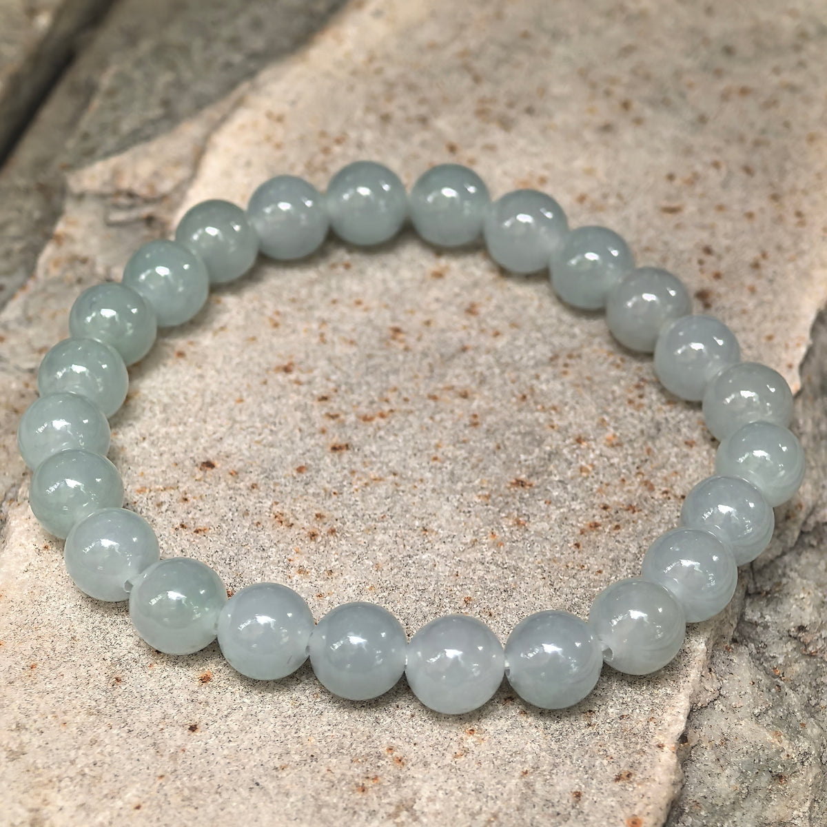 Angled view of a light green jadeite bead bracelet arranged in a single strand on a stone surface.