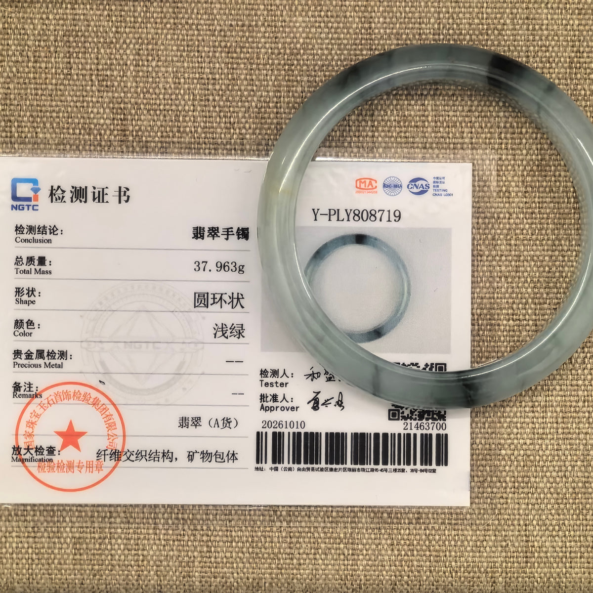 Light green D-shape jadeite bangle with dark floating patterns beside its matching NGTC certificate on a woven fabric surface.