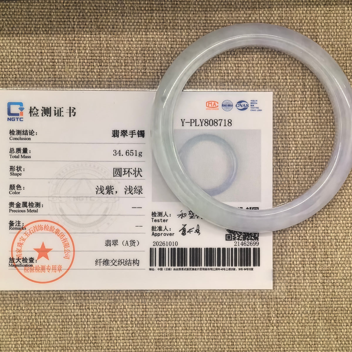 Pale lavender and light green jadeite D-shape bangle beside its matching NGTC certificate on a woven fabric surface.