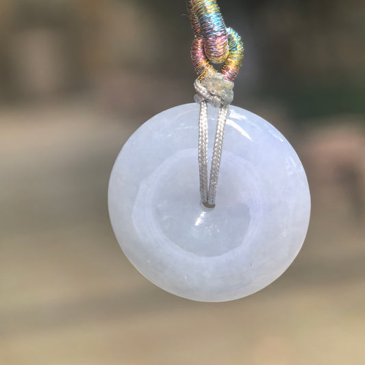 Front view of a pale lavender jadeite Peace Pendant suspended from a gray cord with a multicolored woven knot.