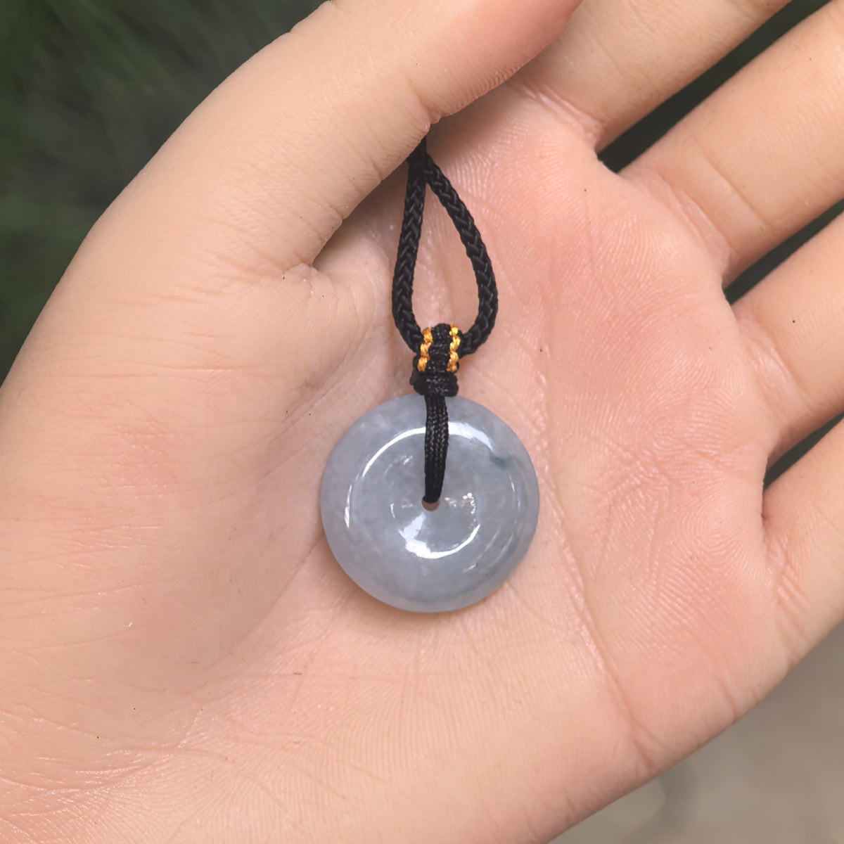 Pale blue jadeite Peace Pendant resting in an open palm, showing its rounded profile, central opening and black braided cord.