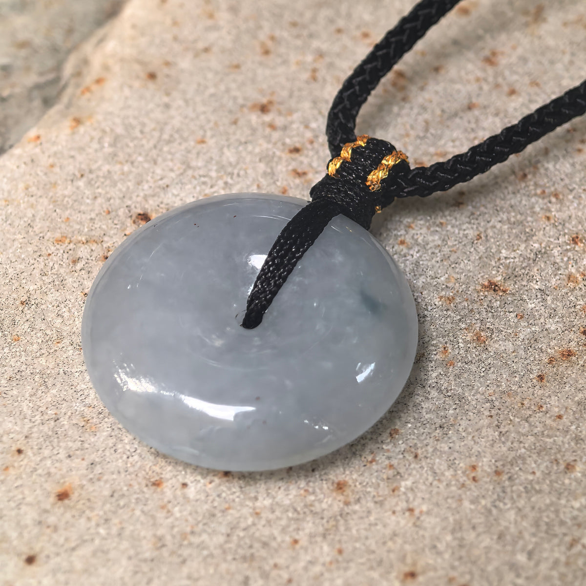 Angled close-up of a pale blue jadeite Peace Pendant on stone, showing its polished face, rounded edge and decorative cord knot.