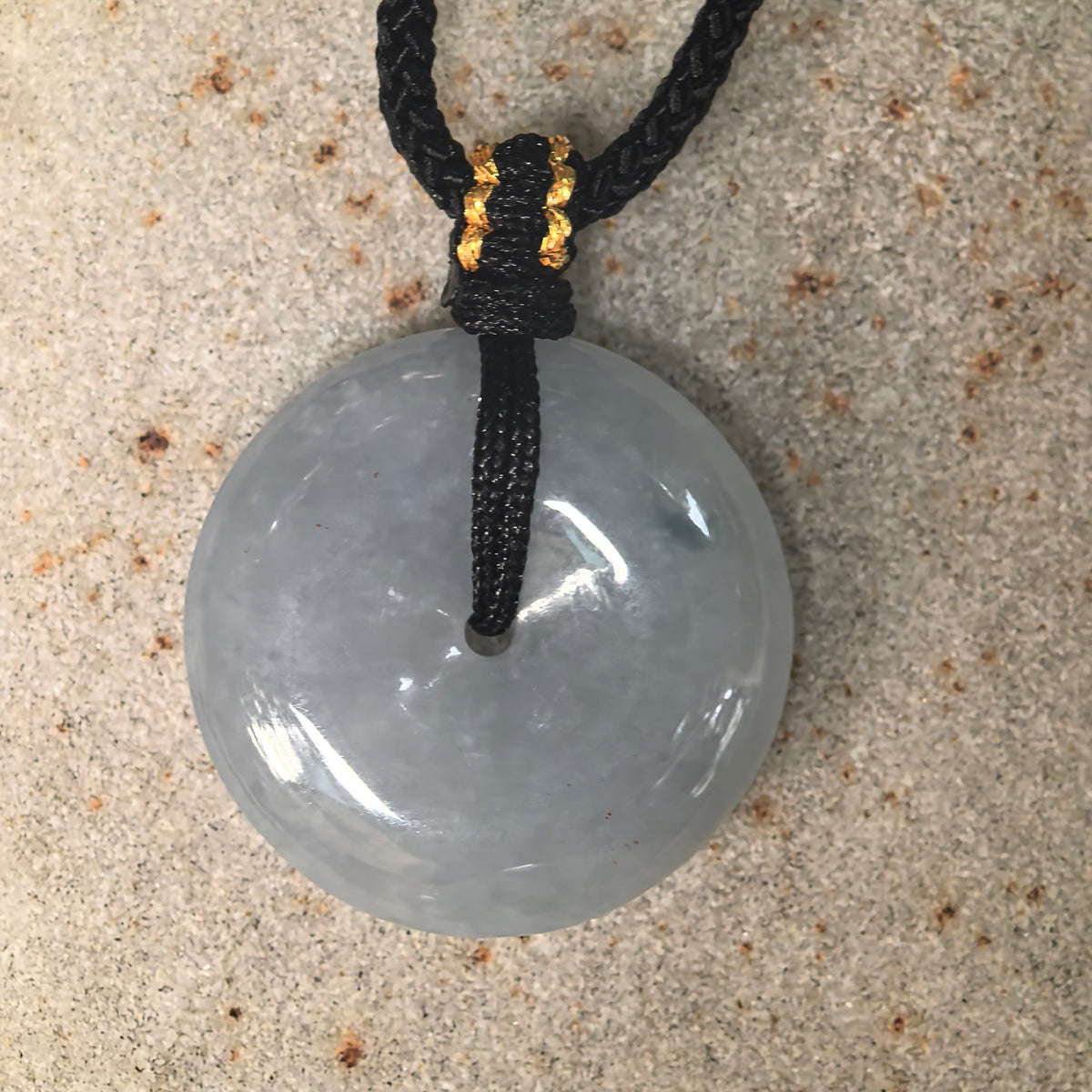 Overhead view of a pale blue jadeite Peace Pendant on textured stone, with a black cord passing through the central opening.