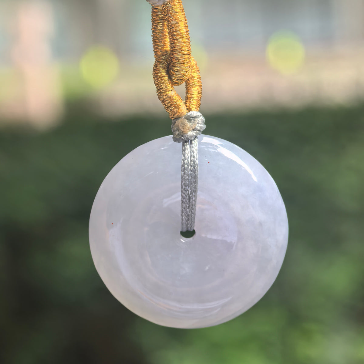 Front view of a pale lavender jadeite Peace Pendant suspended from a knotted cord, showing its circular shape and polished surface.