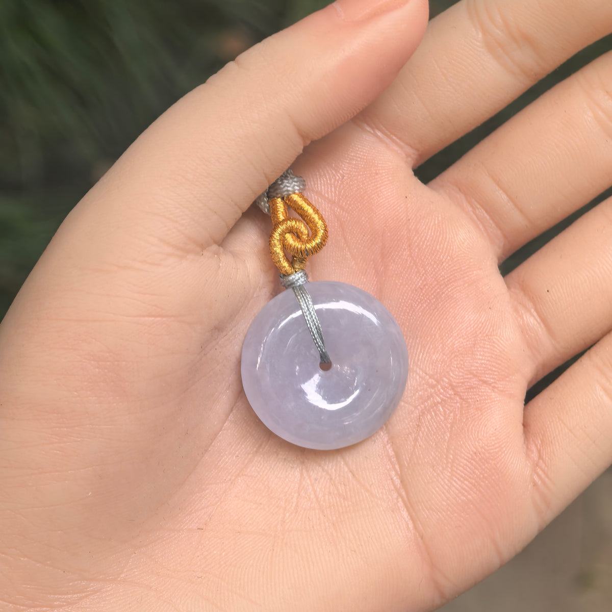 Pale lavender jadeite Peace Pendant resting in an open palm, with a central opening and decorative woven cord.