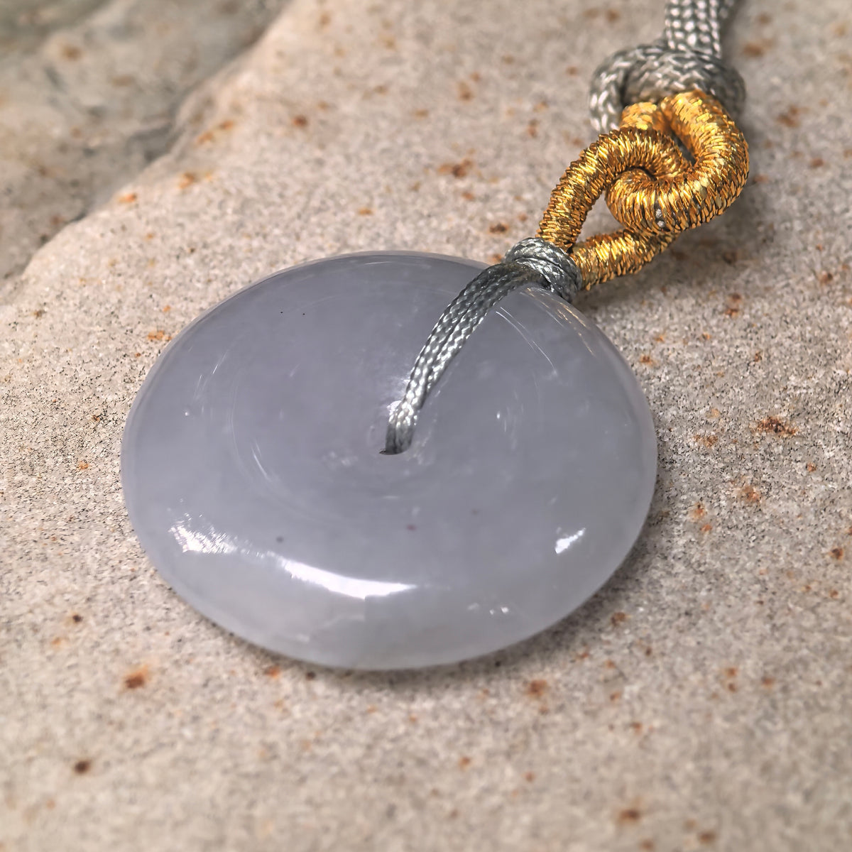 Angled close-up of a lavender jadeite Peace Pendant on stone, showing its rounded edge, polished face and knotted cord.