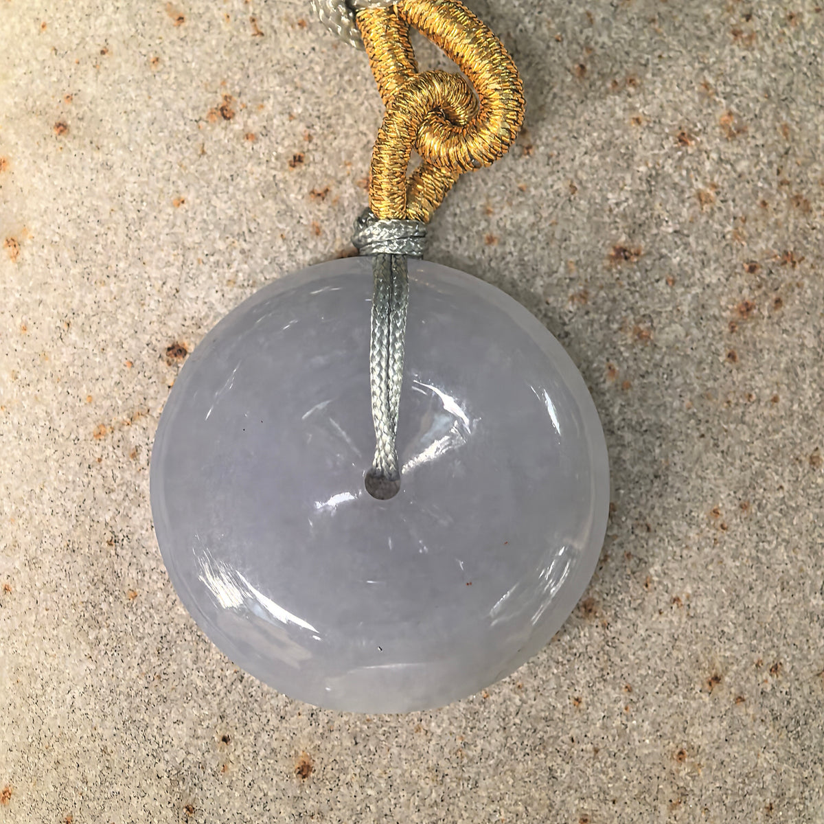 Overhead view of a pale lavender jadeite Peace Pendant on textured stone, showing its circular outline and central opening.