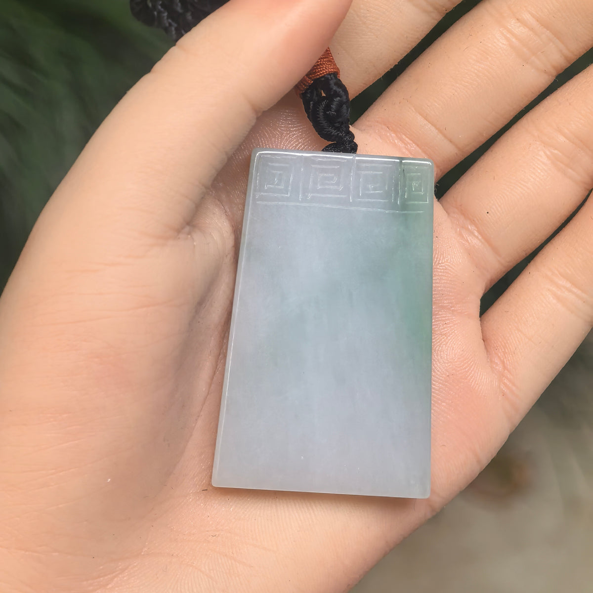 White jadeite Plain Plaque Pendant with green accents resting in an open palm, showing its carved top border and knotted black cord.