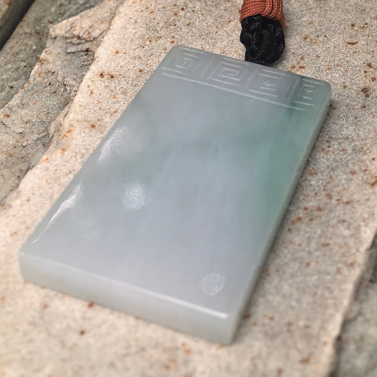 Angled view of the Ruyi-pattern jadeite Plain Plaque Pendant on stone, showing its polished face, green accents and carved upper border.
