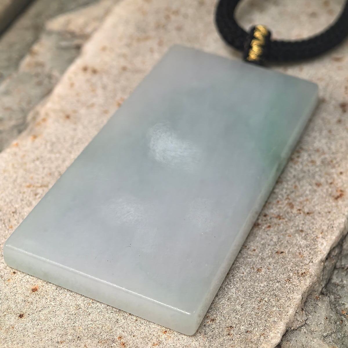 Angled close-up of the uncarved jadeite Plain Plaque Pendant on stone, showing its plain surface, green accents and beveled edge.