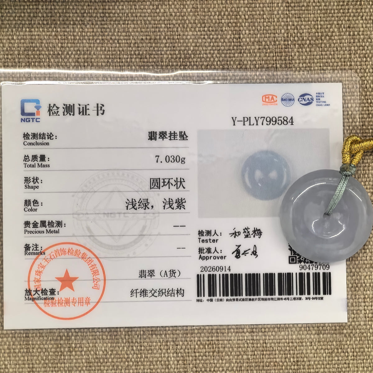 Pale lavender jadeite Peace Pendant with its cord beside the matching NGTC certificate on tan woven fabric.